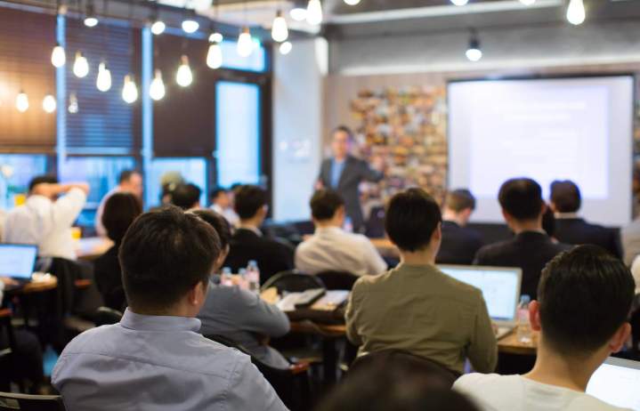 Un orateur présentant un sujet culturel devant une audience attentive, avec des diapositives projetées, dans une salle de conférence moderne.